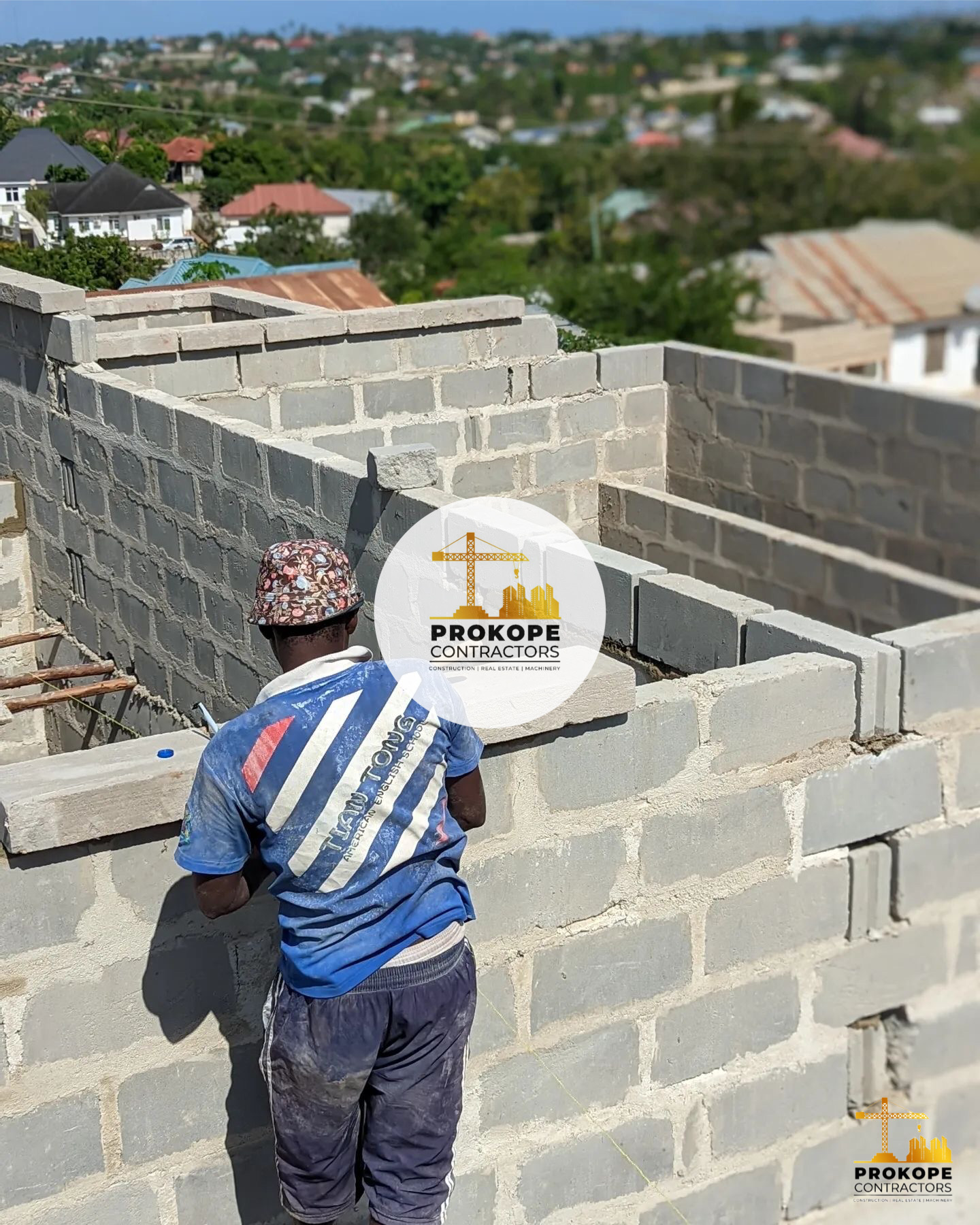 Builder laying blockwork on top of a wall at a construction site with rooftops and greenery in the background